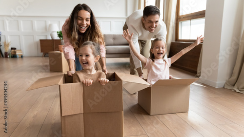Cheerful adorable screaming girls sit inside of big carton boxes during race competition play with parents at new modern home at move day. Bank loan for young family, active games with kids concept