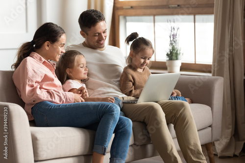 Beautiful full family with two cute daughters resting with laptop seated on sofa. Couple develop offspring watching educational online tv, buy goods on internet, have fun use modern technology concept