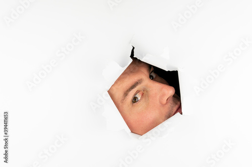 A man looks through a hole in a white wall with one eye.