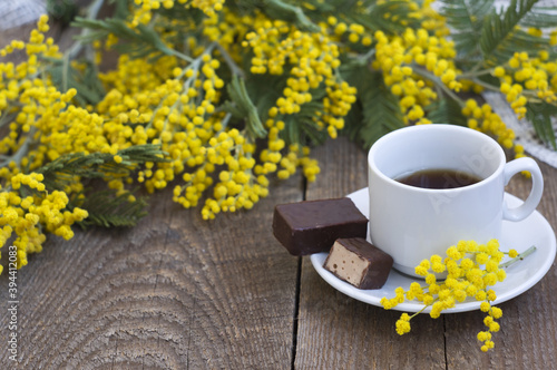 white cup with coffee on plate, candy, yellow flowers mimosa on wooden background