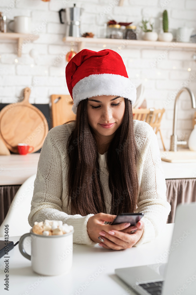 Young brunette woman in santa hat chatting with friends using her mobile phone at the kitchen Young brunette woman in santa hat chatting with friends using her mobile phone at the kitchen