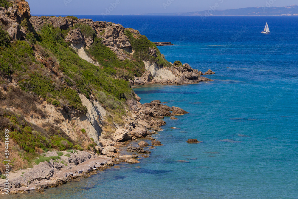 A picturesque seascape - stone shores overgrown with bushes against the backdrop of the blue sea. A lonely sailing yacht is sailing on the water