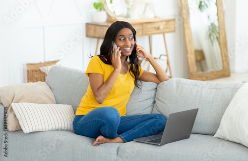 Cheerful young black woman chatting on smartphone and using laptop computer on couch at home