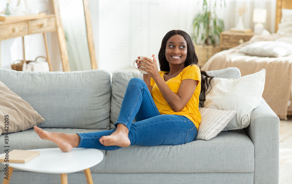 Obraz premium Lovely young black woman having coffee break on comfy couch at home, copy space