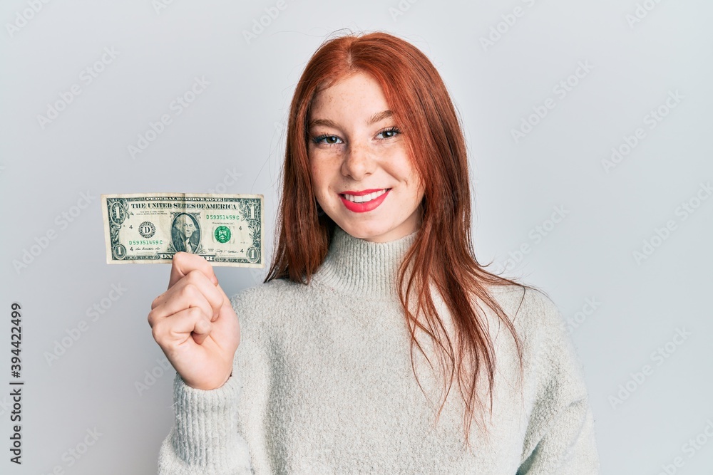Young red head girl holding 1 dollar banknote looking positive and happy standing and smiling with a confident smile showing teeth Young red head girl holding 1 dollar banknote looking positive and happy standing and smiling with a confident smile showing teeth