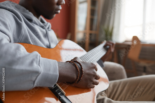 Close up of young African-American man playing acoustic guitar while sitting on couch and making music at home, copy space