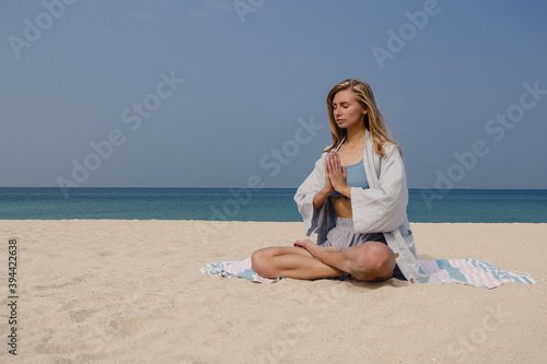 Blonde young girl practices yoga and meditation in the lotus position on the beach on a sunny day.