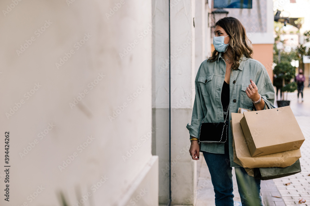 Portrait of a white woman in a green jacket, black bag and mask looking at a store window while holding many paper bags. Portrait of a white woman in a green jacket, black bag and mask looking at a store window while holding many paper bags.
