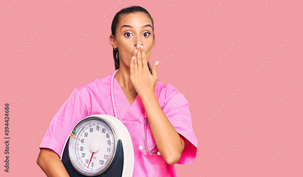 Young hispanic woman as nutritionist doctor holding weighing machine covering mouth with hand, shocked and afraid for mistake. surprised expression Young hispanic woman as nutritionist doctor holding weighing machine covering mouth with hand, shocked and afraid for mistake. surprised expression