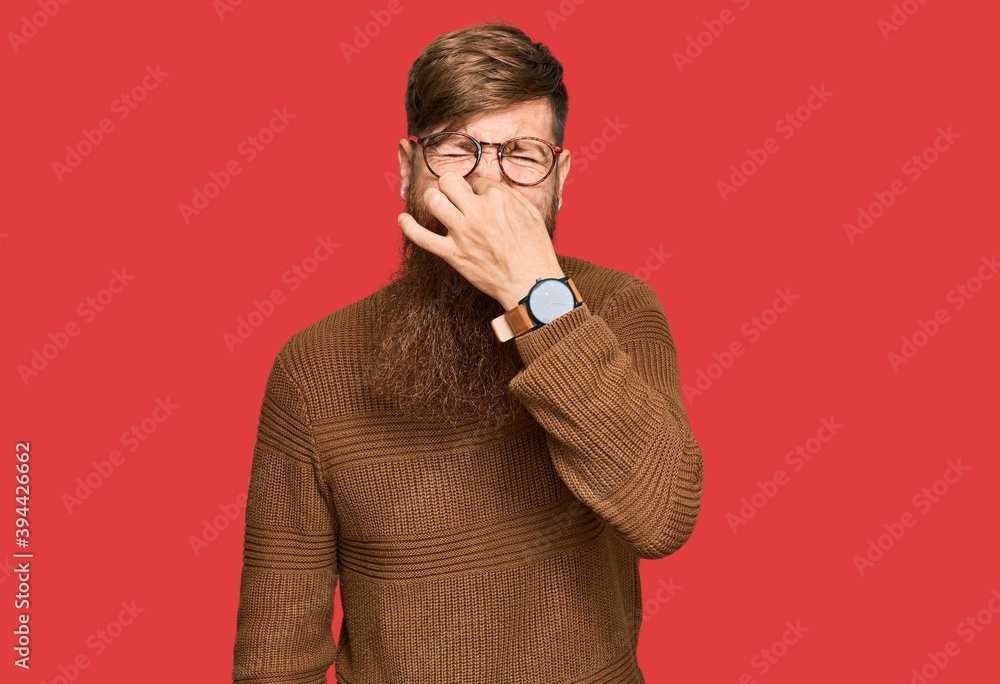 Young irish redhead man wearing casual clothes and glasses smelling something stinky and disgusting, intolerable smell, holding breath with fingers on nose. bad smell Young irish redhead man wearing casual clothes and glasses smelling something stinky and disgusting, intolerable smell, holding breath with fingers on nose. bad smell