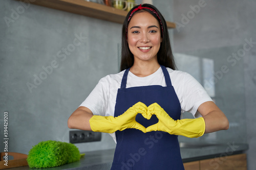 Love to Clean. Cheerful young woman, cleaning lady wearing protective gloves, smiling at camera, showing heart sign while standing in the kitchen, ready for cleaning the house
