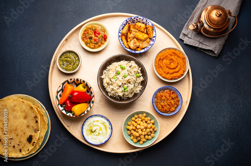 Indian Thali - selection of various dishes served on round wooden platter. Assorted Indian vegetarian meze with boiled basmati rice, paneer, dal, chutney, chapati bread and brass teapot. Top view