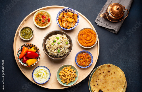 Indian Thali - selection of various dishes served on round wooden platter. Assorted Indian vegetarian meze with boiled basmati rice, paneer, dal, chutney, chapati bread and brass teapot. Top view