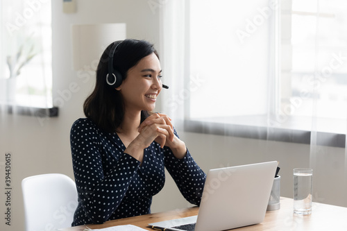 Smiling Asian businesswoman wearing headset using laptop, looking to aside, dreaming about new job opportunities, happy woman student watching webinar, listening to lecture, studying, home office