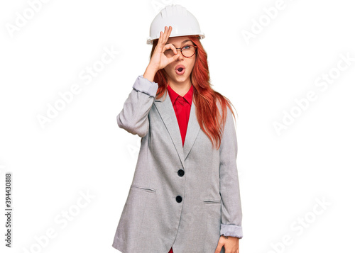 Young redhead woman wearing architect hardhat doing ok gesture shocked with surprised face, eye looking through fingers. unbelieving expression.