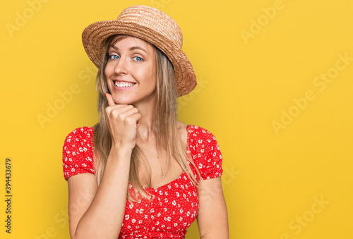 Beautiful caucasian woman wearing summer hat looking confident at the camera with smile with crossed arms and hand raised on chin. thinking positive.