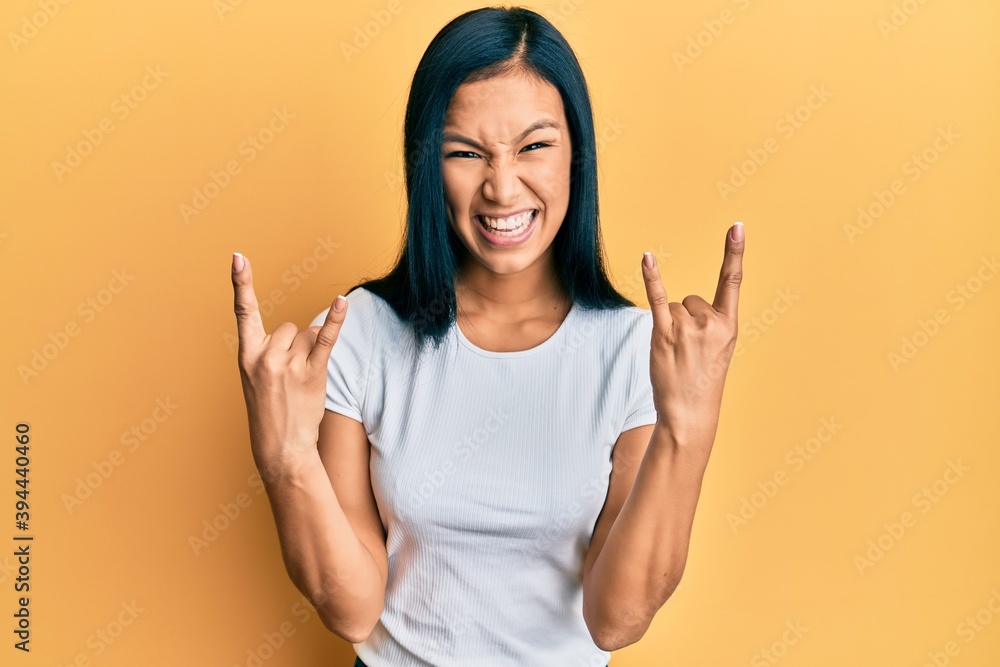 Beautiful hispanic woman wearing casual white tshirt shouting with crazy expression doing rock symbol with hands up. music star. heavy concept. Beautiful hispanic woman wearing casual white tshirt shouting with crazy expression doing rock symbol with hands up. music star. heavy concept.