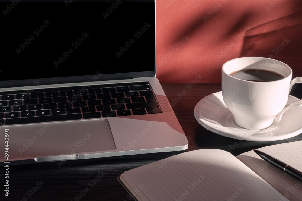 Laptop, cup of coffee, notepad and pen in the morning light with shadows. Home office desktop. Educational and business concept. Laptop, cup of coffee, notepad and pen in the morning light with shadows. Home office desktop. Educational and business concept.