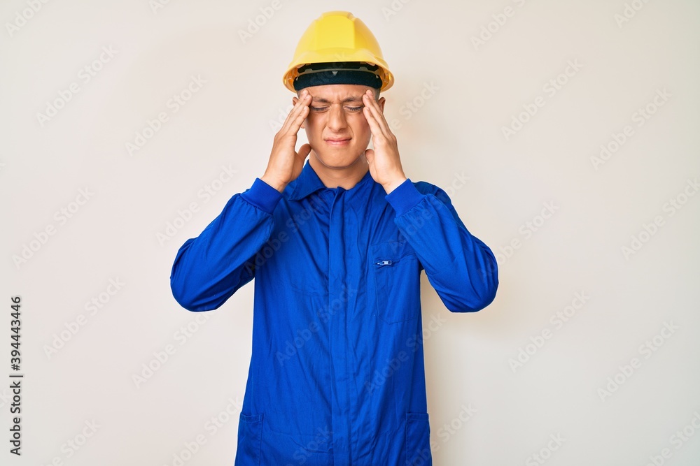 Young hispanic boy wearing worker uniform and hardhat suffering from headache desperate and stressed because pain and migraine. hands on head.