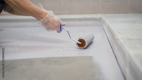 A worker paints the concrete floor with white paint. A worker paints the floor with a roller. Manual painting of a white floor with a paint roller for waterproofing