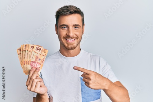 Handsome caucasian man holding mexican pesos smiling happy pointing with hand and finger photo