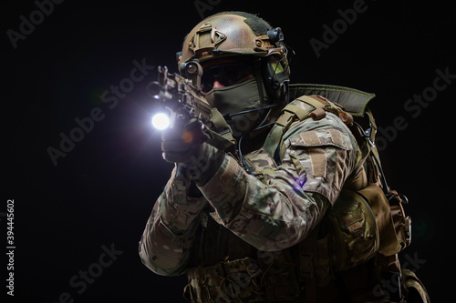 Close up portrait of army soldier in military camouflage uniform, helmet, glasses and face hidden behind mask holds a rifle near face and takes aim using an optical sight isolated on black background © satyrenko