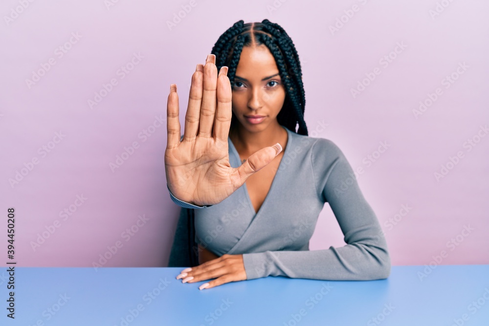 Beautiful hispanic woman wearing casual clothes sitting on the table doing stop sing with palm of the hand. warning expression with negative and serious gesture on the face. Beautiful hispanic woman wearing casual clothes sitting on the table doing stop sing with palm of the hand. warning expression with negative and serious gesture on the face.