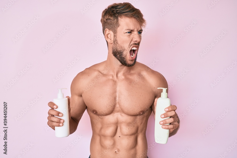 Fototapeta premium Young caucasian man shirtless holding cream lotion angry and mad screaming frustrated and furious, shouting with anger. rage and aggressive concept.