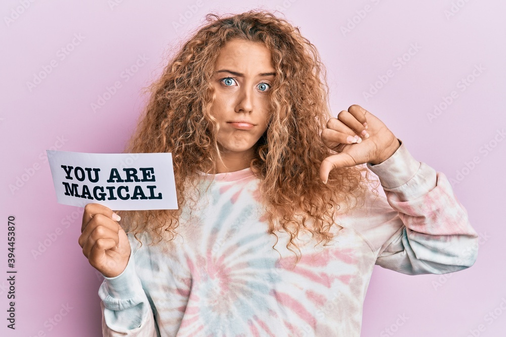 Beautiful caucasian teenager girl holding you are magical banner with angry face, negative sign showing dislike with thumbs down, rejection concept Beautiful caucasian teenager girl holding you are magical banner with angry face, negative sign showing dislike with thumbs down, rejection concept