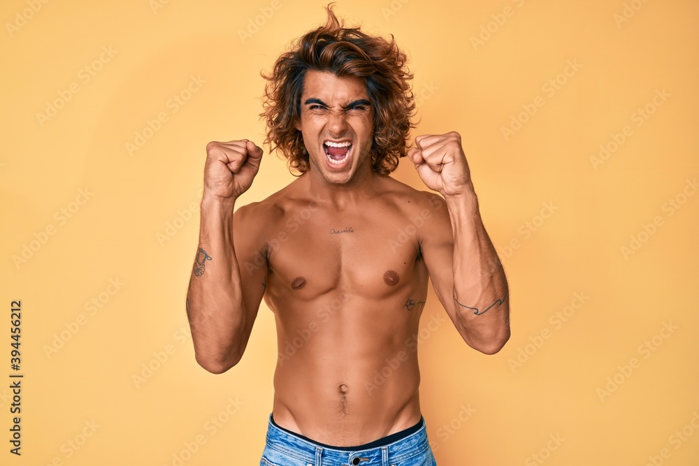 Young hispanic man standing shirtless angry and mad raising fists frustrated and furious while shouting with anger. rage and aggressive concept.