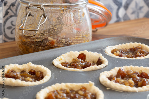 Mince pies in a baking tray with mincemeat in a clip glass jar and a spoon.  On a wood worktop with a tiled background