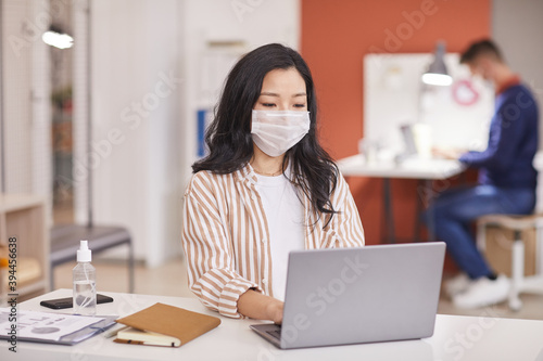 Portrait of young Asian woman wearing mask and using laptop while working at desk in office with bottle of sanitizer in foreground, copy space