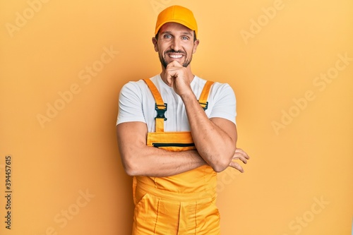 Young handsome man wearing handyman uniform over yellow background looking confident at the camera smiling with crossed arms and hand raised on chin. thinking positive.