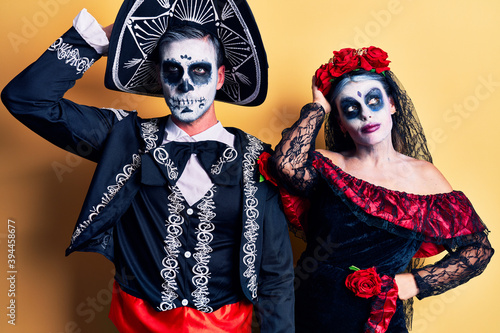Young couple wearing mexican day of the dead costume over yellow confuse and wondering about question. uncertain with doubt, thinking with hand on head. pensive concept.