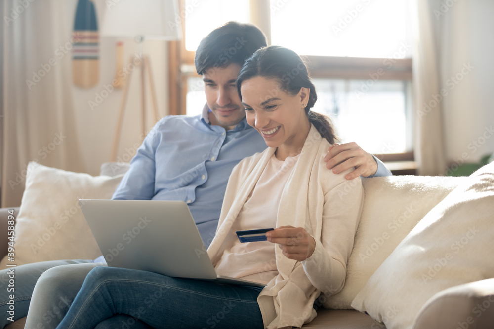 Home shopping. Happy millennial family couple hugging on cozy couch at living room sitting by laptop pc holding plastic bank card preparing to make payment purchase online using e-banking service app Home shopping. Happy millennial family couple hugging on cozy couch at living room sitting by laptop pc holding plastic bank card preparing to make payment purchase online using e-banking service app