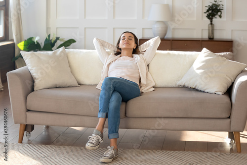 Calm and positive. Happy serene young female relaxing on couch at living room leaning back on pillows, taking break of household chores, breathing fresh air, dreaming about spending weekend vacation