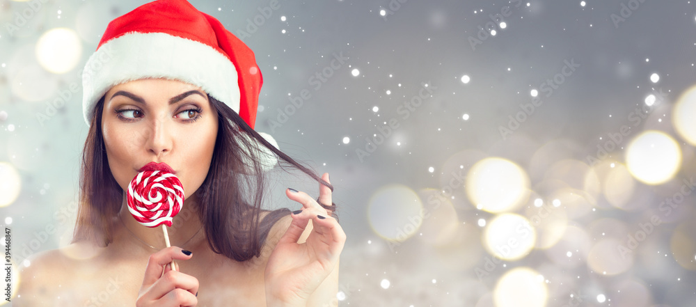 Christmas woman. Beauty model girl in Santa Claus hat with red lips and xmas lollipop candy. Closeup portrait on holiday gray background