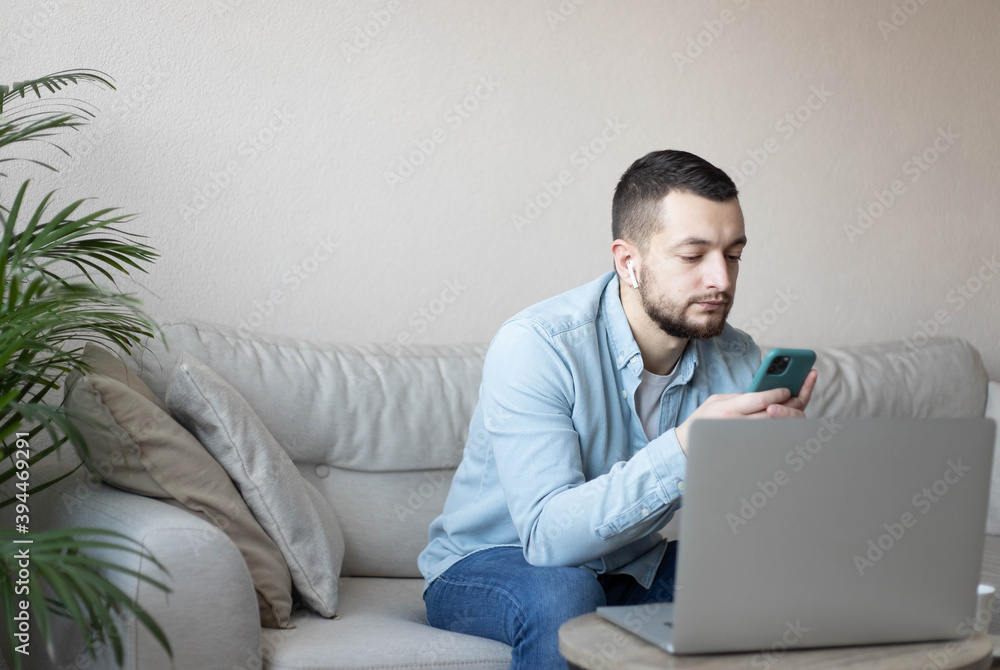 Young man reading sms message on smartphone sitting with laptop device.Smiling young guy typing on mobile phone while sitting on a sofa at home with notebook. Young man reading sms message on smartphone sitting with laptop device.Smiling young guy typing on mobile phone while sitting on a sofa at home with notebook.