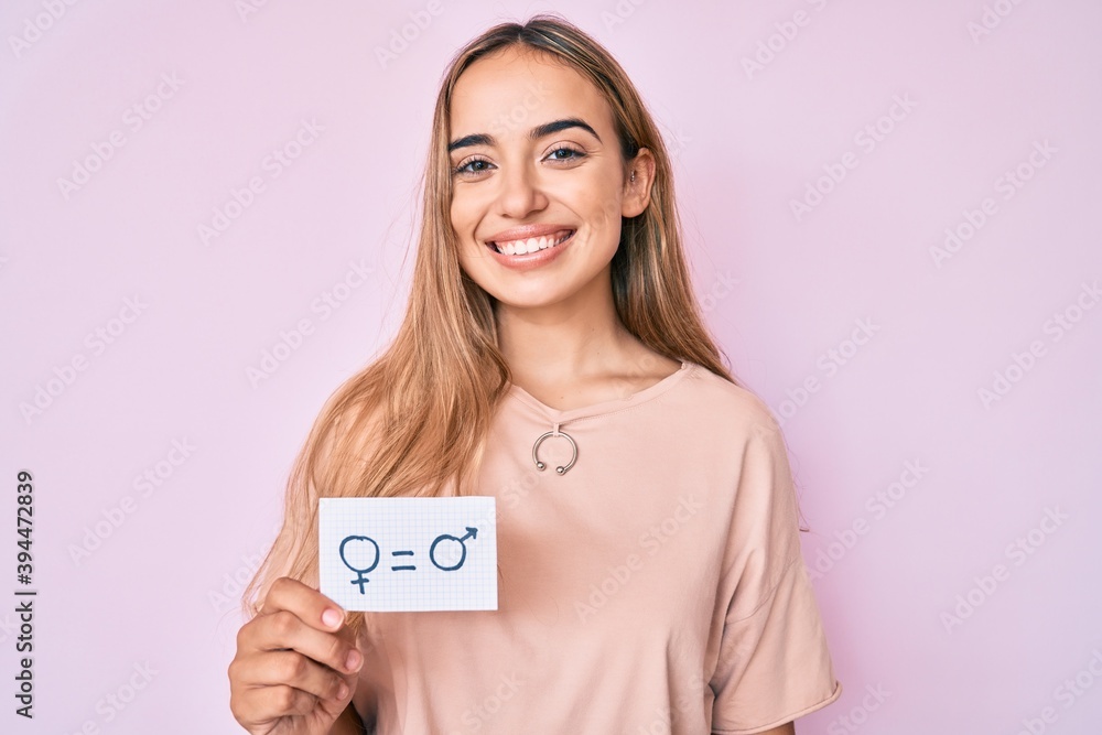 Young beautiful blonde woman holding we are equal paper looking positive and happy standing and smiling with a confident smile showing teeth Young beautiful blonde woman holding we are equal paper looking positive and happy standing and smiling with a confident smile showing teeth