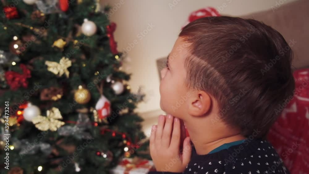 Dark-haired boy at home looking at the Christmas tree, enjoying the view. The boy prays . Celebrating Christmas at home. Dark-haired boy at home looking at the Christmas tree, enjoying the view. The boy prays . Celebrating Christmas at home.