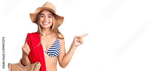 Young beautiful blonde woman wearing bikini and hat holding summer wicker handbag smiling happy pointing with hand and finger to the side photo