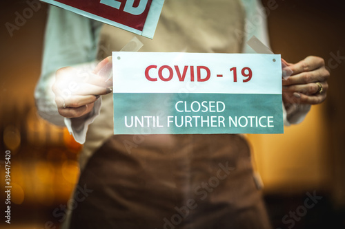 Female silhouette with a beige apron, standing behind the glass door with the sign ''closed'' and putting the covid19 notice