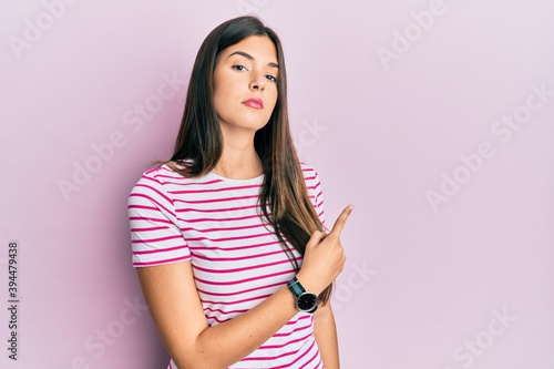 Young brunette woman wearing casual clothes over pink background pointing with hand finger to the side showing advertisement, serious and calm face © Krakenimages.com