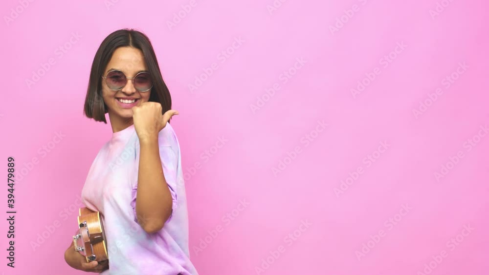 Young latin woman playing ukelele points with thumb finger away, laughing and carefree Young latin woman playing ukelele points with thumb finger away, laughing and carefree