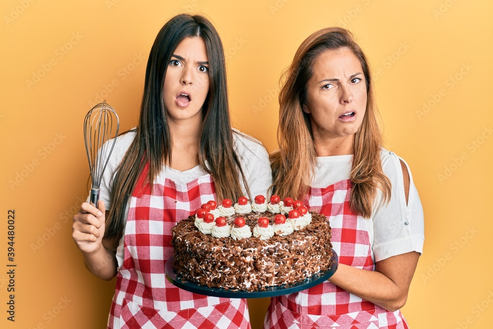 Hispanic family of mother and daughter wearing baker apron holding homemade cake in shock face, looking skeptical and sarcastic, surprised with open mouth