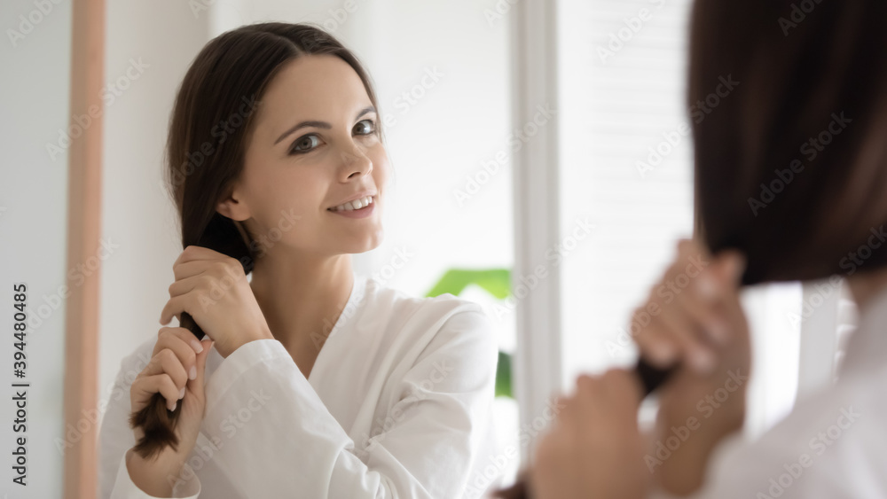 Strong and beautiful. Smiling young lady looking at mirror checking hair condition after treatment course, feeling content with its thick shiny healthy look, creating new stylish hairdo after shower Strong and beautiful. Smiling young lady looking at mirror checking hair condition after treatment course, feeling content with its thick shiny healthy look, creating new stylish hairdo after shower