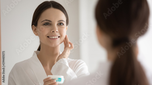 Amazing effect. Excited overjoyed young woman looking at mirror touching healthy soft smooth velvet skin on cheek after using cosmetic cream from professional beauty care line, delighted with result