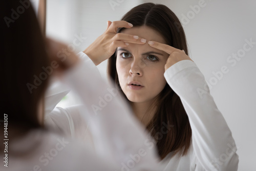 Oh God, I have a zit. Worried anxious young woman teenager standing by mirror annoyed of bad skin condition acne rash irritation dermatitis allergic response to new face care cream cosmetic product