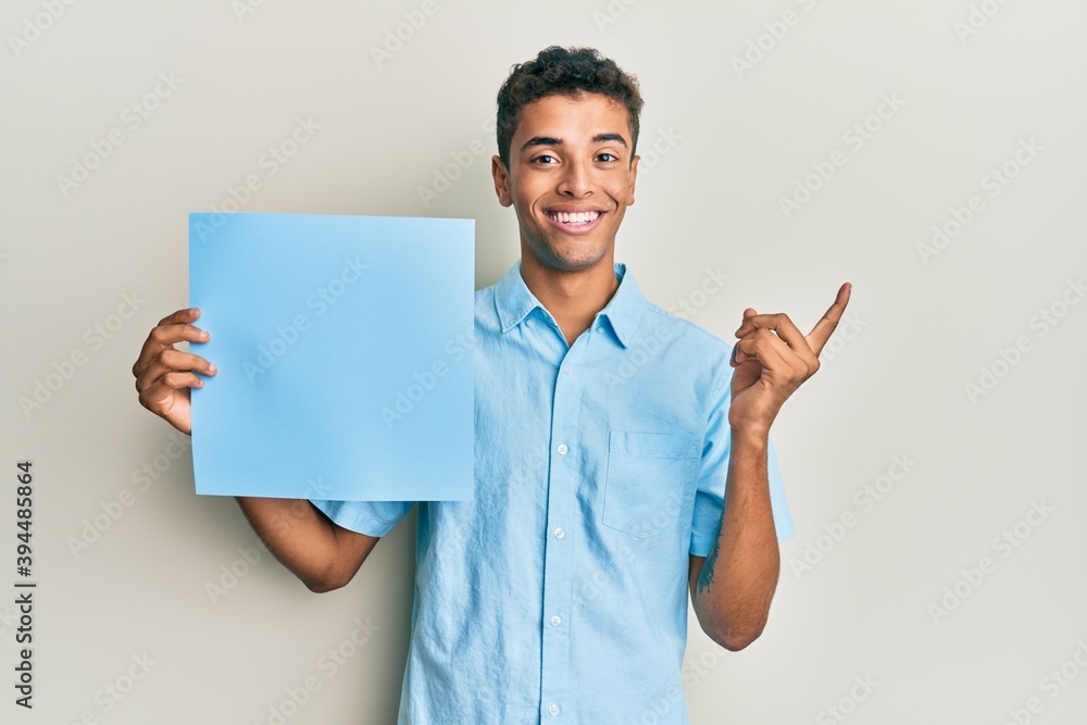Young handsome african american man holding blue blank empty banner smiling happy pointing with hand and finger to the side Young handsome african american man holding blue blank empty banner smiling happy pointing with hand and finger to the side