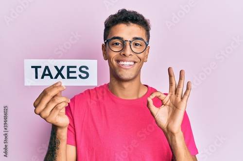 Young handsome african american man holding taxes word doing ok sign with fingers, smiling friendly gesturing excellent symbol © Krakenimages.com